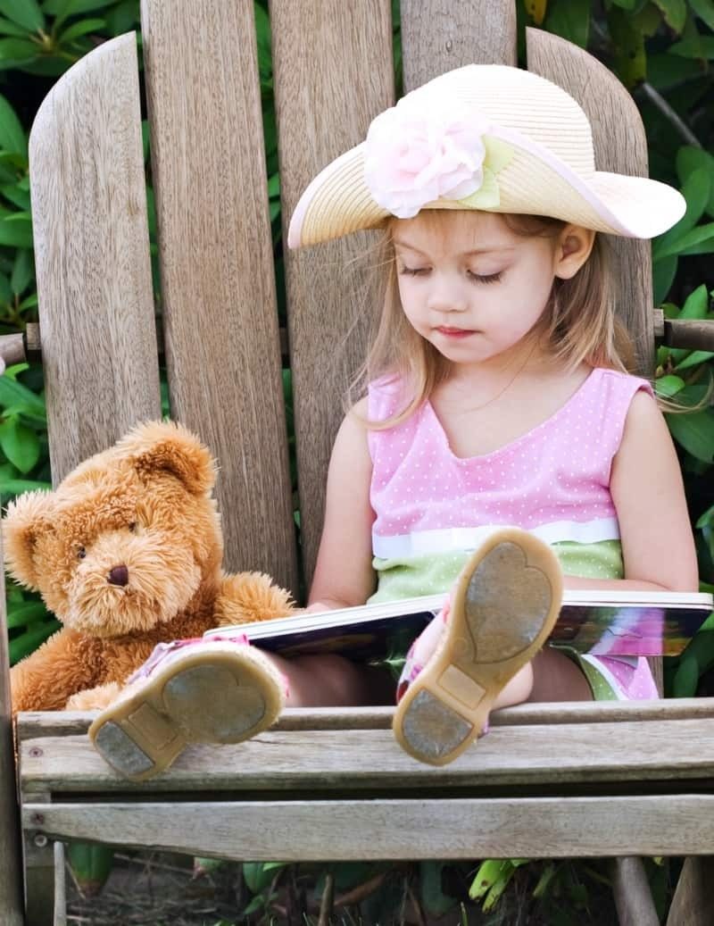 Young girl in a sun hat reading a picture book next to her teddy bear